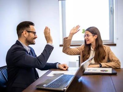 Un hombre y una mujer en una oficina celebrando con un "high five" frente a una laptop y gráficos financieros, simbolizando decisiones estratégicas exitosas.