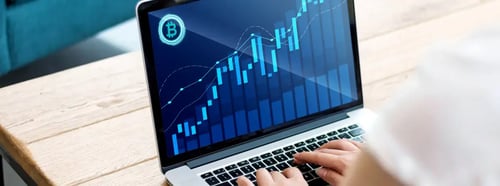 Persona utilizando una laptop con gráfico financiero de velas y líneas ascendentes en pantalla, con símbolo de Bitcoin, representando análisis de criptomonedas.