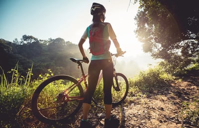 Mujer ciclista con mochila observando el amanecer en un sendero rodeado de vegetación, junto a su bicicleta de montaña.