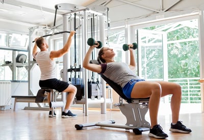 Hombre y mujer ejercitándose en gimnasio, utilizando pesas y máquina de poleas.
