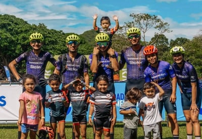 Grupo de ciclistas con cascos de colores y niños posando para una foto en un evento deportivo al aire libre.