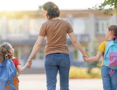Mujer de espaldas caminando con dos niñas hacia la escuela, tomadas de la mano.
