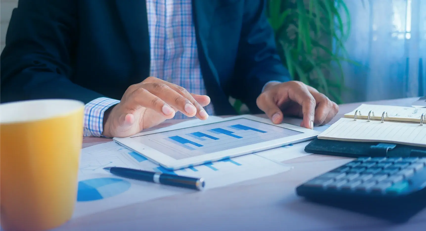 Persona de negocios analizando gráficos financieros en una tablet sobre un escritorio con documentos, calculadora y agenda.