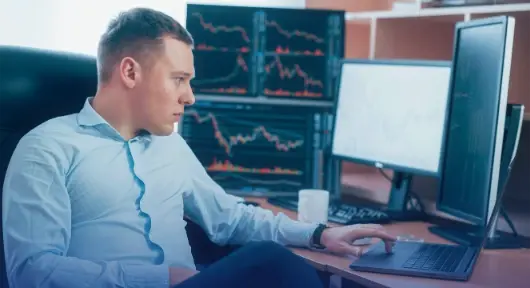 Hombre observa gráficas en múltiples pantallas mientras analiza datos financieros en su oficina.