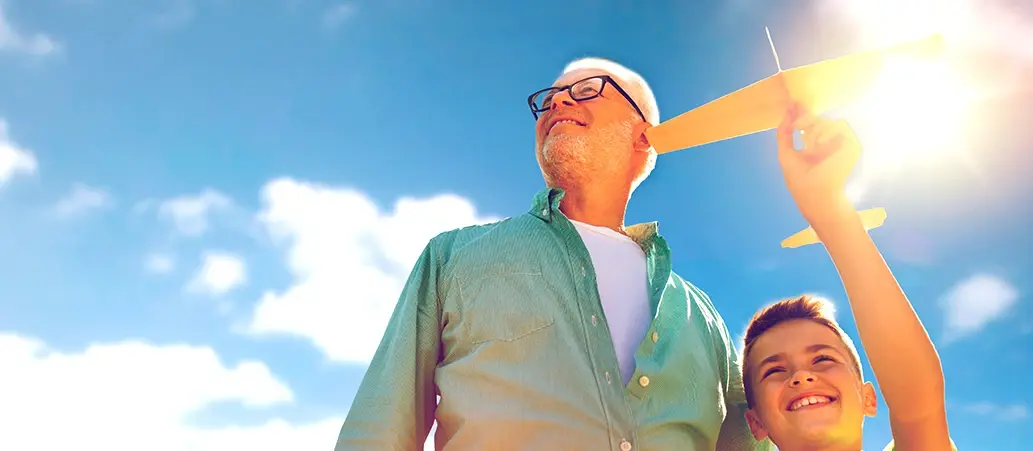 Hombre mayor con gafas de sol y camisa verde junto a un niño que sostiene un avión de juguete, con cielo azul y sol de fondo