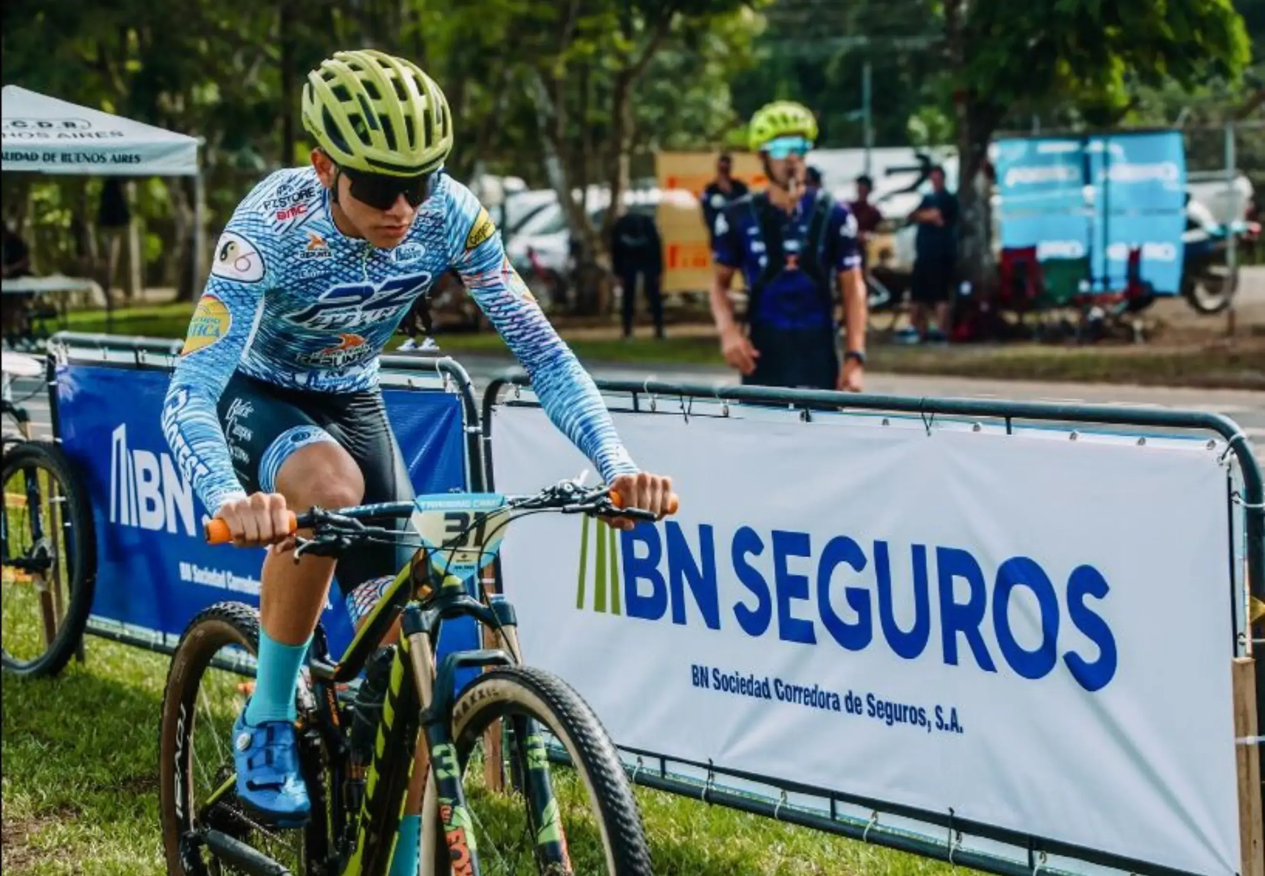 Ciclista con casco amarillo y gafas oscuras participando en una competencia de ciclismo de montaña, con un rótulo publicitario de BN Seguros al fondo.