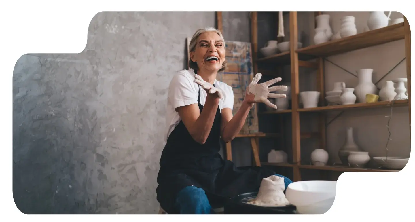 Banner de blog que muestra a una mujer mayor riendo con las manos cubiertas de arcilla en un taller de alfarería.