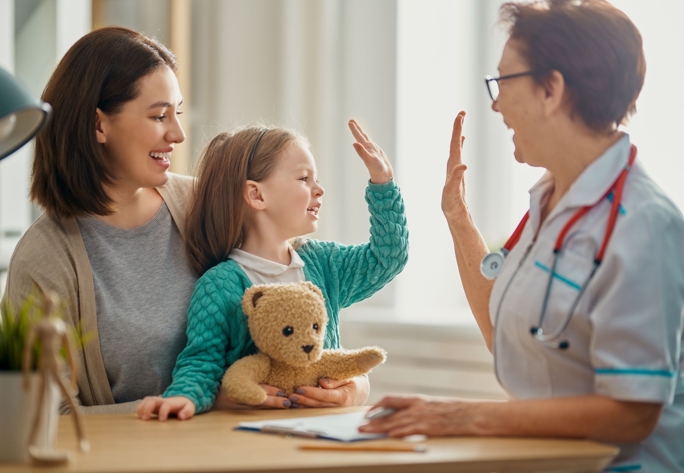 Niña con osito de peluche dando un ‘high five’ a doctora mientras su madre sonríe.