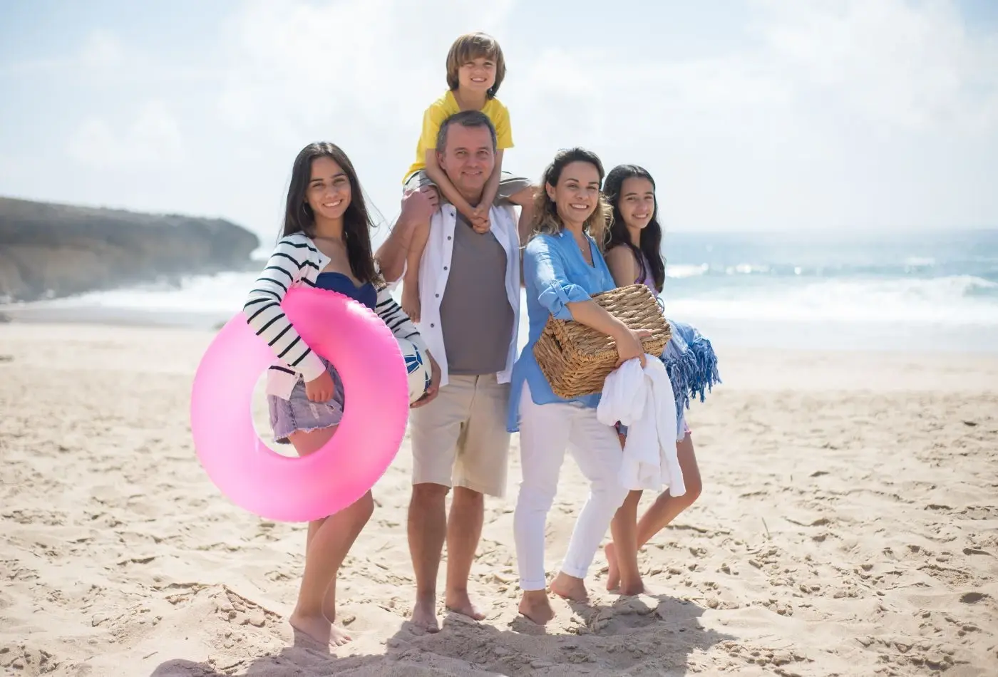 Familia en la playa sonriendo, con flotador rosa y cesta de picnic, disfrutando de un día soleado junto al mar.