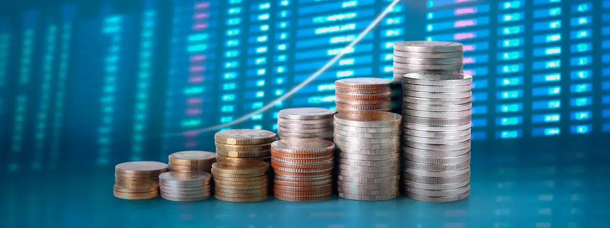 Montones de monedas apiladas en orden ascendente sobre una superficie azul, con una pantalla digital de fondo que muestra cifras y gráficos financieros, simbolizando crecimiento económico.