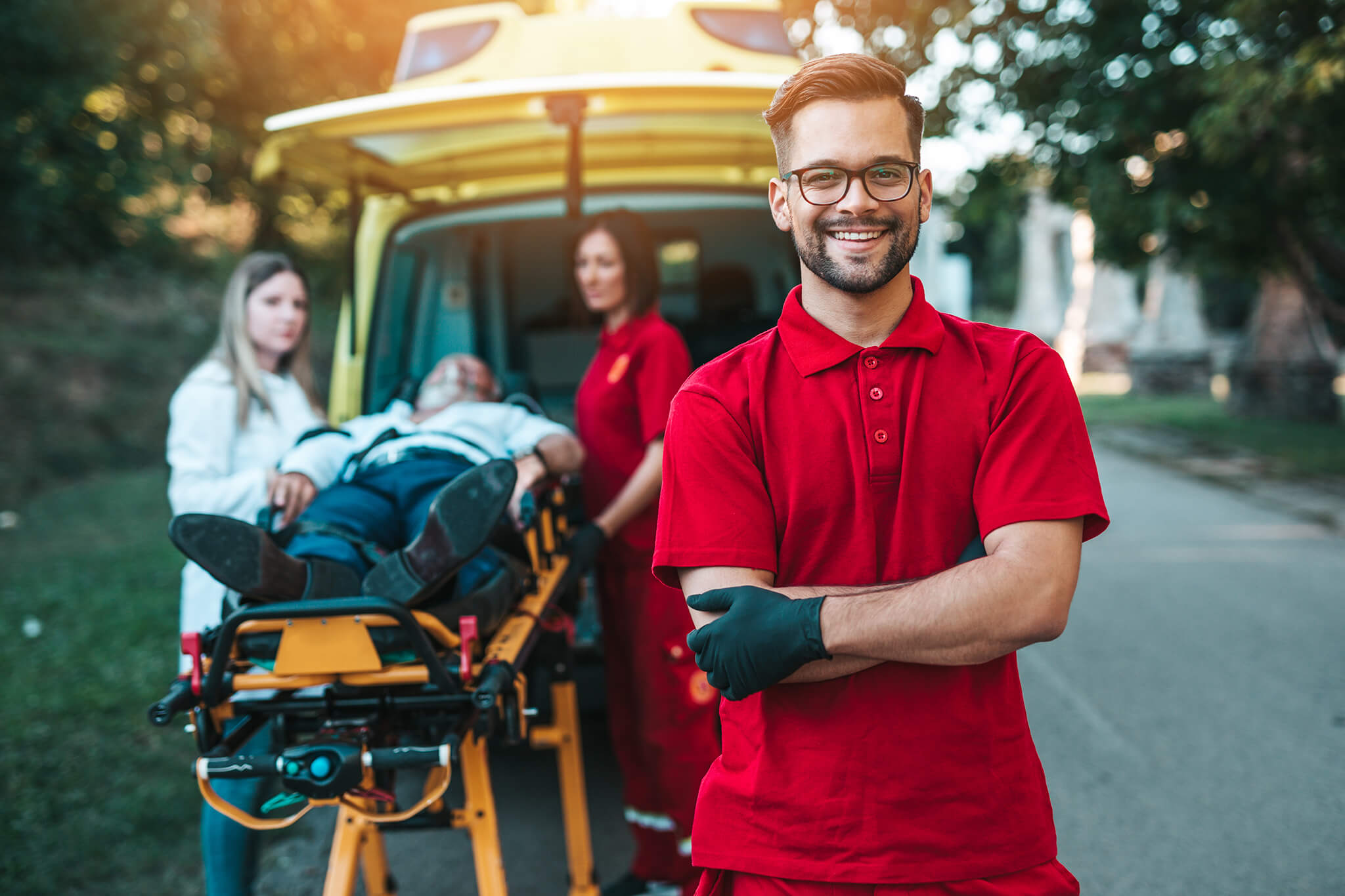 Paramédico sonriendo frente a una ambulancia mientras compañeros atienden a un paciente en camilla.