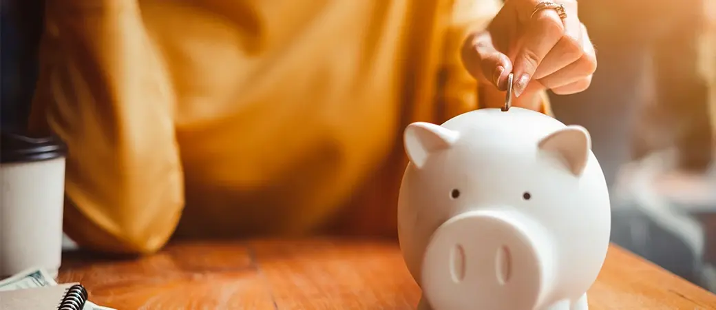 Persona insertando una moneda en una alcancía blanca con forma de cerdito sobre una mesa de madera