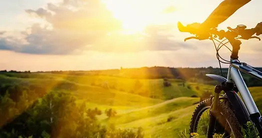 Bicicleta en primer plano durante un atardecer en el campo, con colinas verdes y rayos de sol iluminando el paisaje.