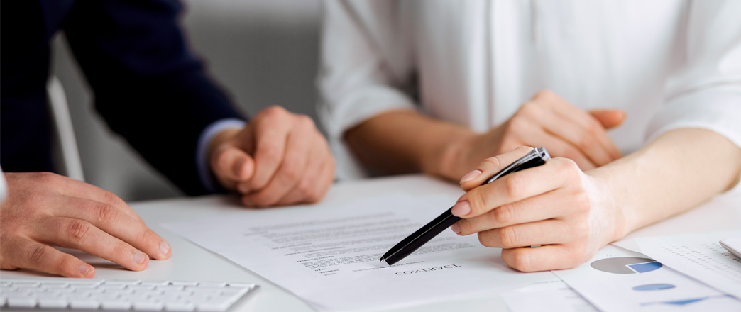 Persona señalando un documento con un bolígrafo mientras conversa con otra persona en un entorno de oficina.