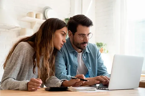 Pareja joven revisando información financiera en una laptop desde la cocina de su hogar, con calculadora y bolígrafo sobre la mesa.
