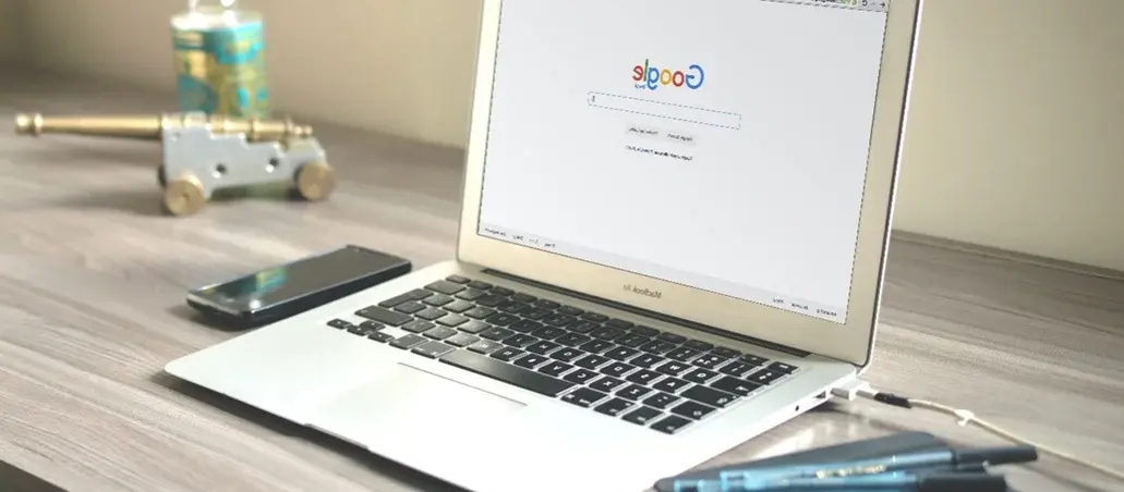 Computadora portátil sobre escritorio con buscador de Google en pantalla, junto a un teléfono móvil y bolígrafos.