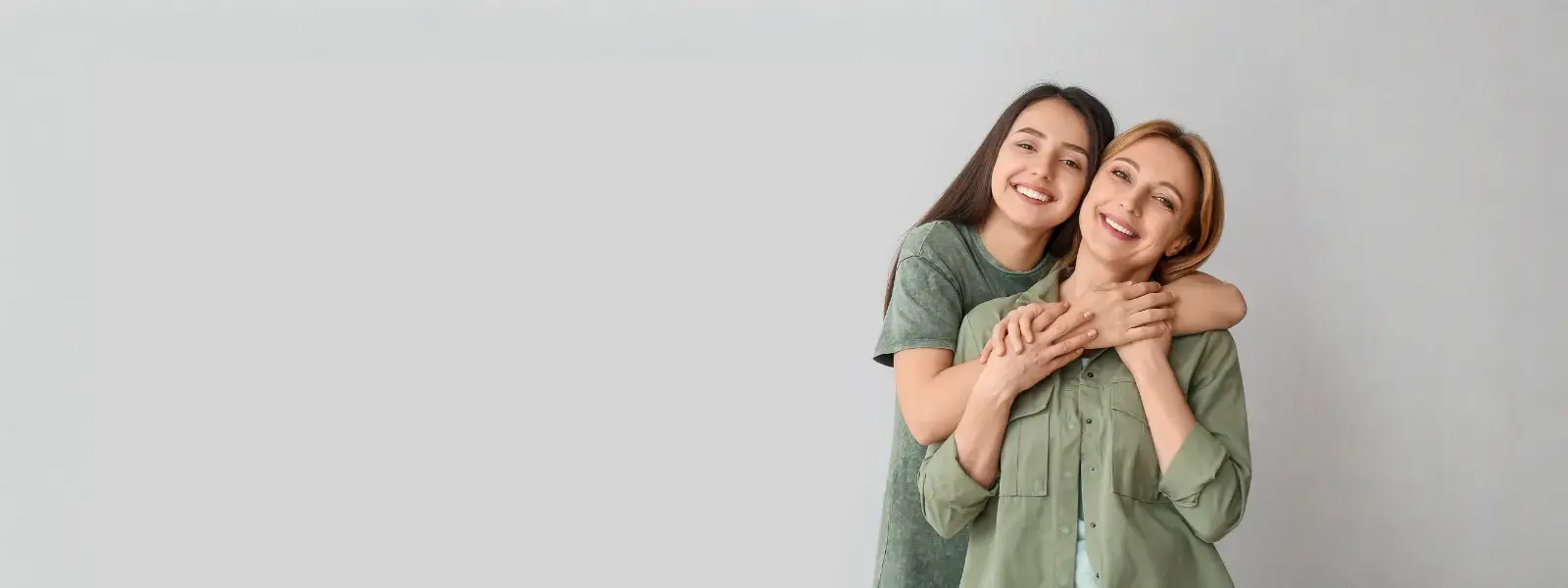 Una madre y su hija adolescente, ambas vestidas con camisas de color verde oliva, posan sonrientes y abrazadas frente a un fondo gris claro, reflejando unión y cariño familiar.