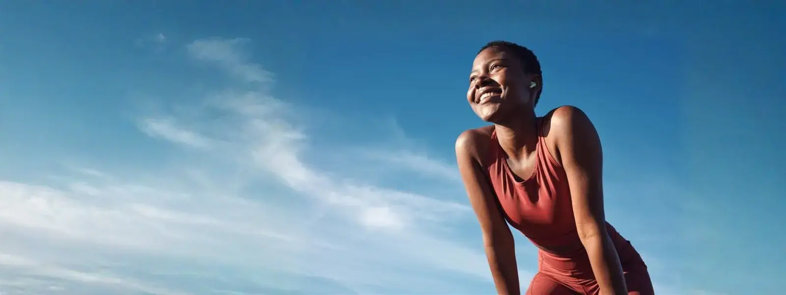 Una mujer con ropa deportiva de color terracota sonríe mientras descansa y mira hacia arriba, bajo un cielo azul brillante con nubes ligeras, transmitiendo una sensación de salud y energía.