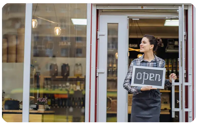 Mujer con delantal sosteniendo cartel de ‘open’ en la entrada de su tienda.