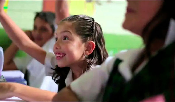 Niña en uniforme escolar sonriendo y levantando la mano en un aula.
