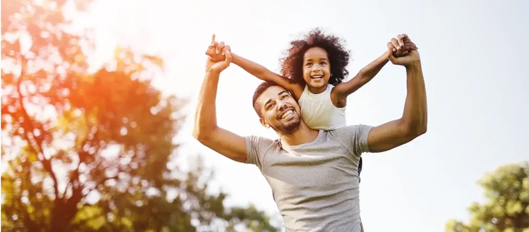 Padre cargando en hombros a su hija pequeña mientras sonríen en un parque al aire libre.