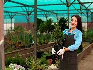 Mujer sonriente sosteniendo una planta en una maceta dentro de un vivero.