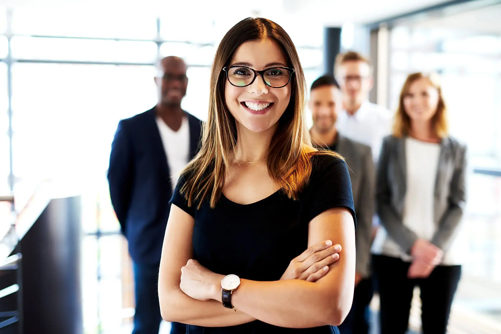 Mujer joven con gafas sonriendo y brazos cruzados, con equipo de trabajo desenfocado al fondo.